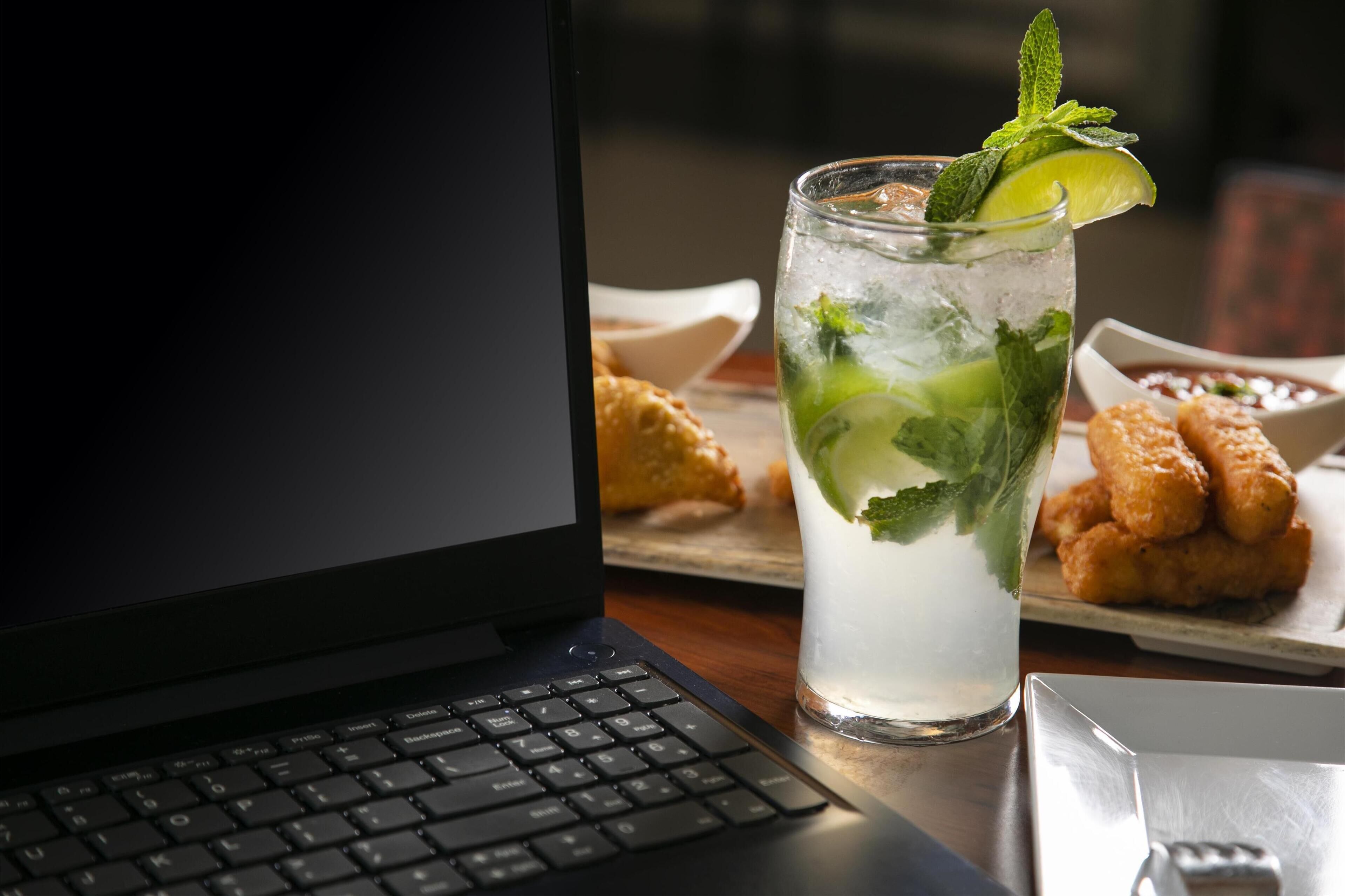 Una computadora portátil sobre un escritorio, con un vaso de bebida helada adornado con menta y lima, y un plato de comida frita al fondo.