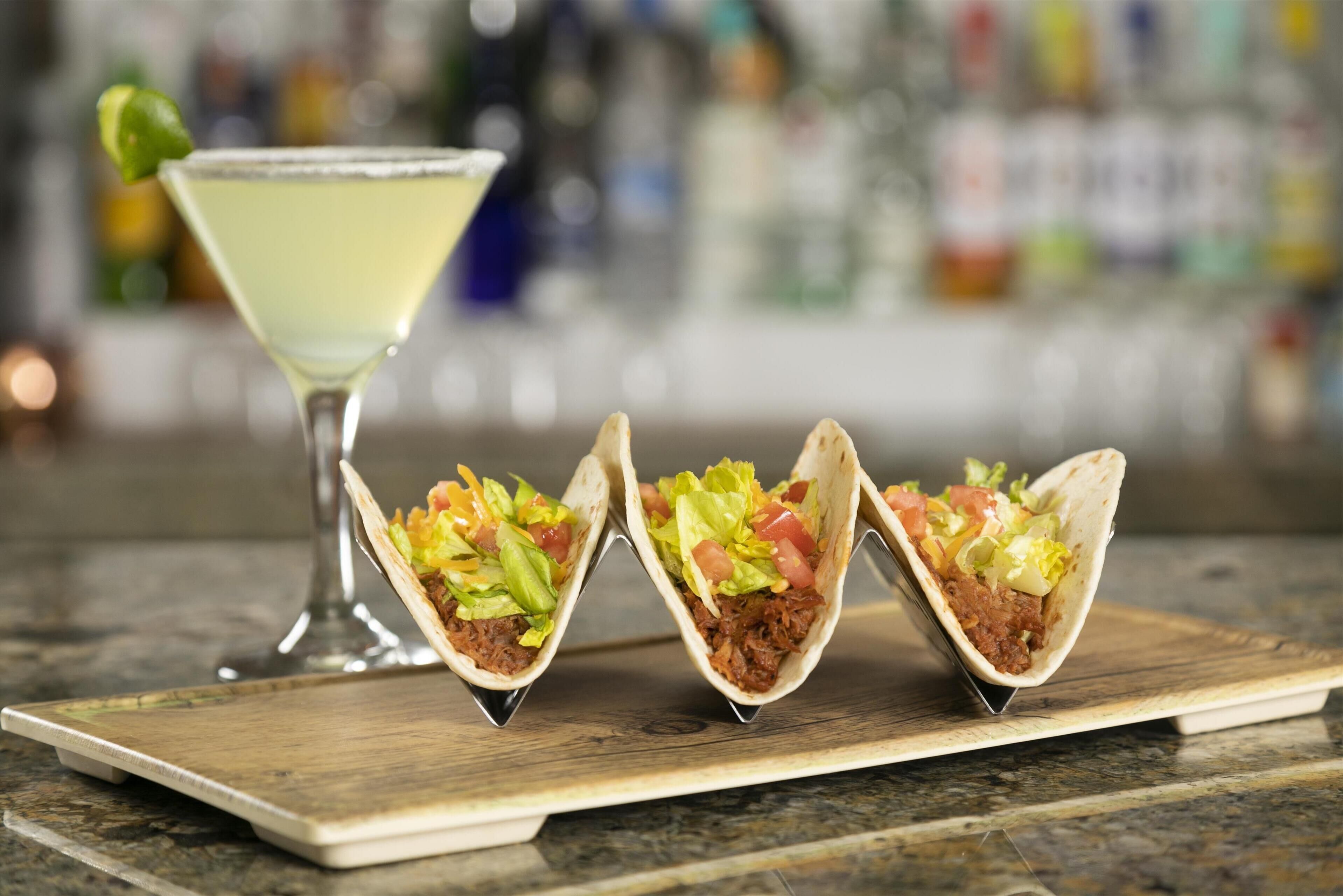 Tre tacos på en träbricka, med ett martiniglas i bakgrunden.