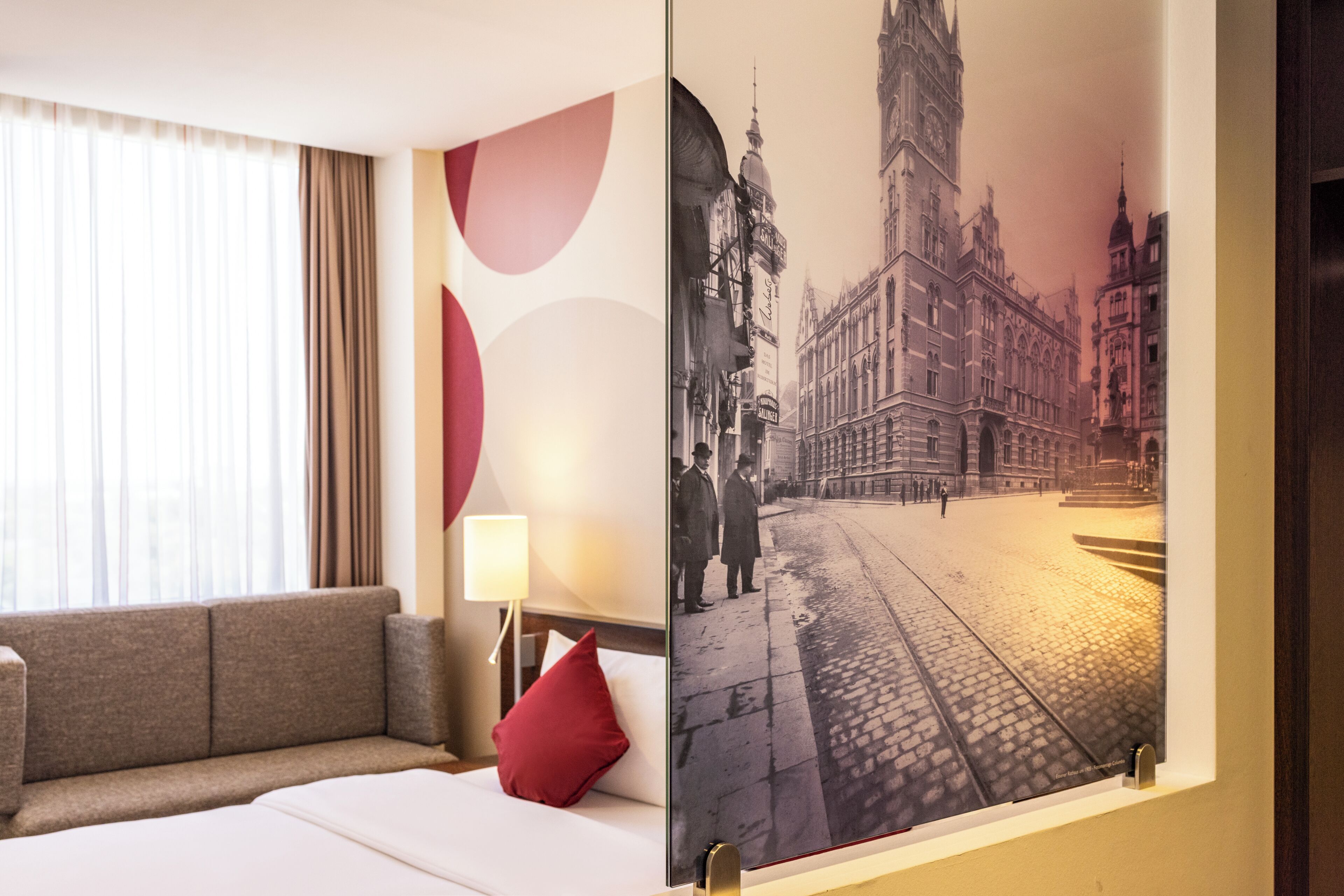 A hotel room with a bed, a red pillow, a nightstand with a lamp, and a wall with a large historical photograph.