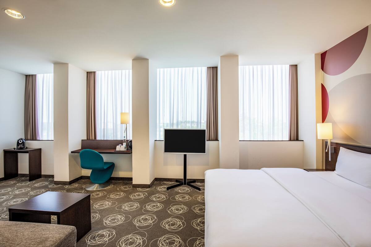 Une chambre d’hôtel équipée d’un lit, d’un bureau, d’une chaise, d’une télévision et de grandes fenêtres avec des rideaux.