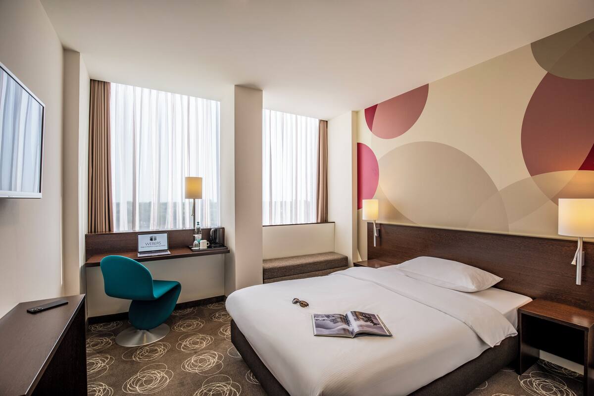 Une chambre d’hôtel avec un lit, un bureau avec un ordinateur, une chaise et une fenêtre avec des rideaux.