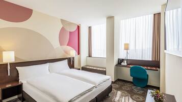 A hotel room with a double bed, a desk, a chair, and a window with curtains.