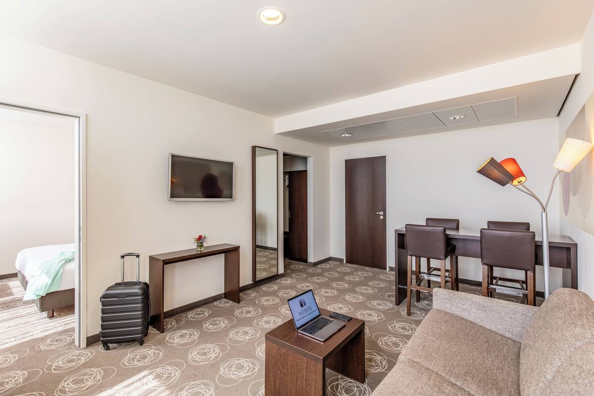 Une chambre d’hôtel comprenant un lit, un bureau, un ordinateur portable, une valise, un canapé, une table à manger avec des chaises et une lampe.