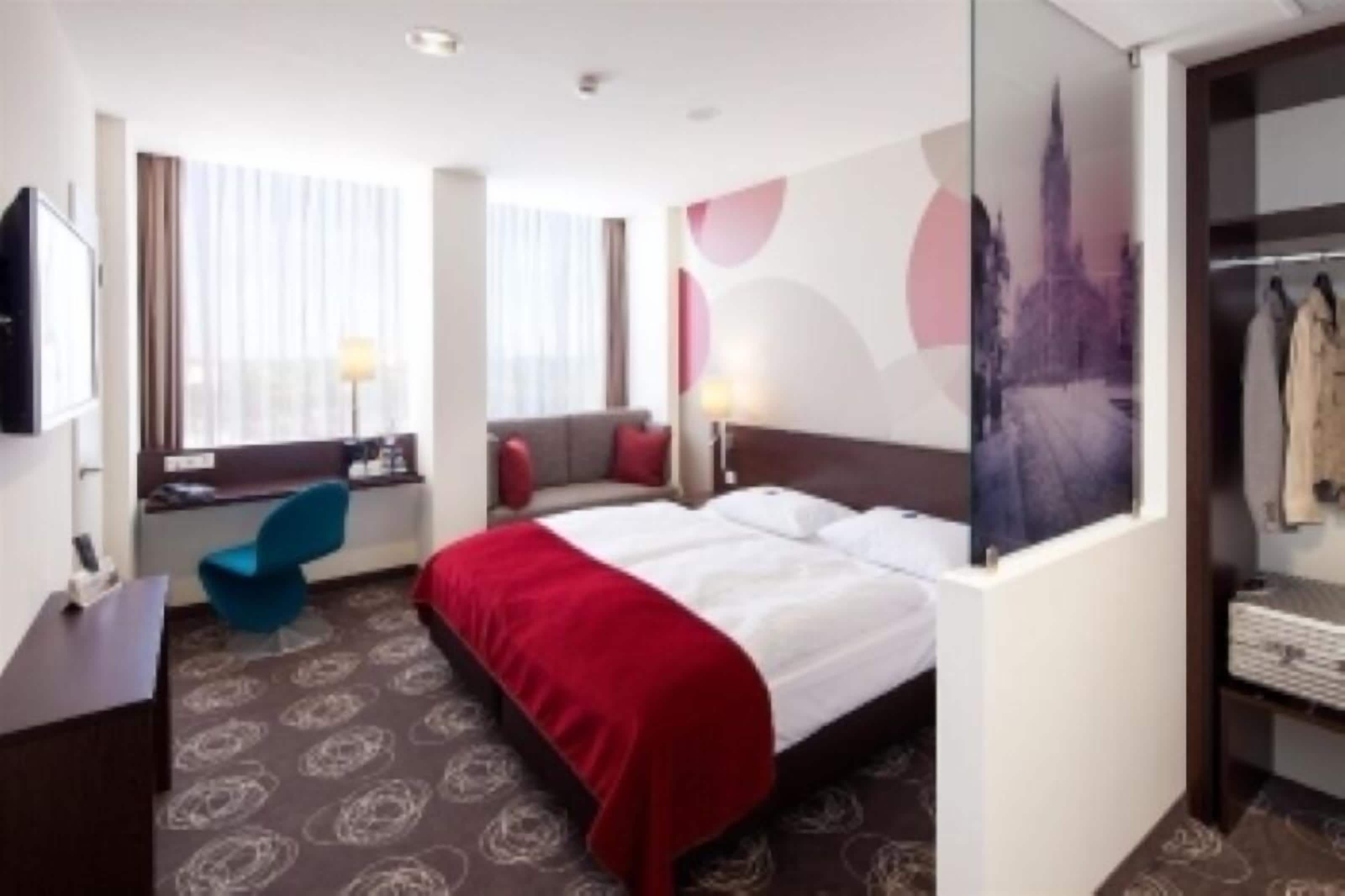 Ein Hotelzimmer mit Bett, Schreibtisch, Stuhl, Kleiderschrank und einem fenster mit Vorhängen.