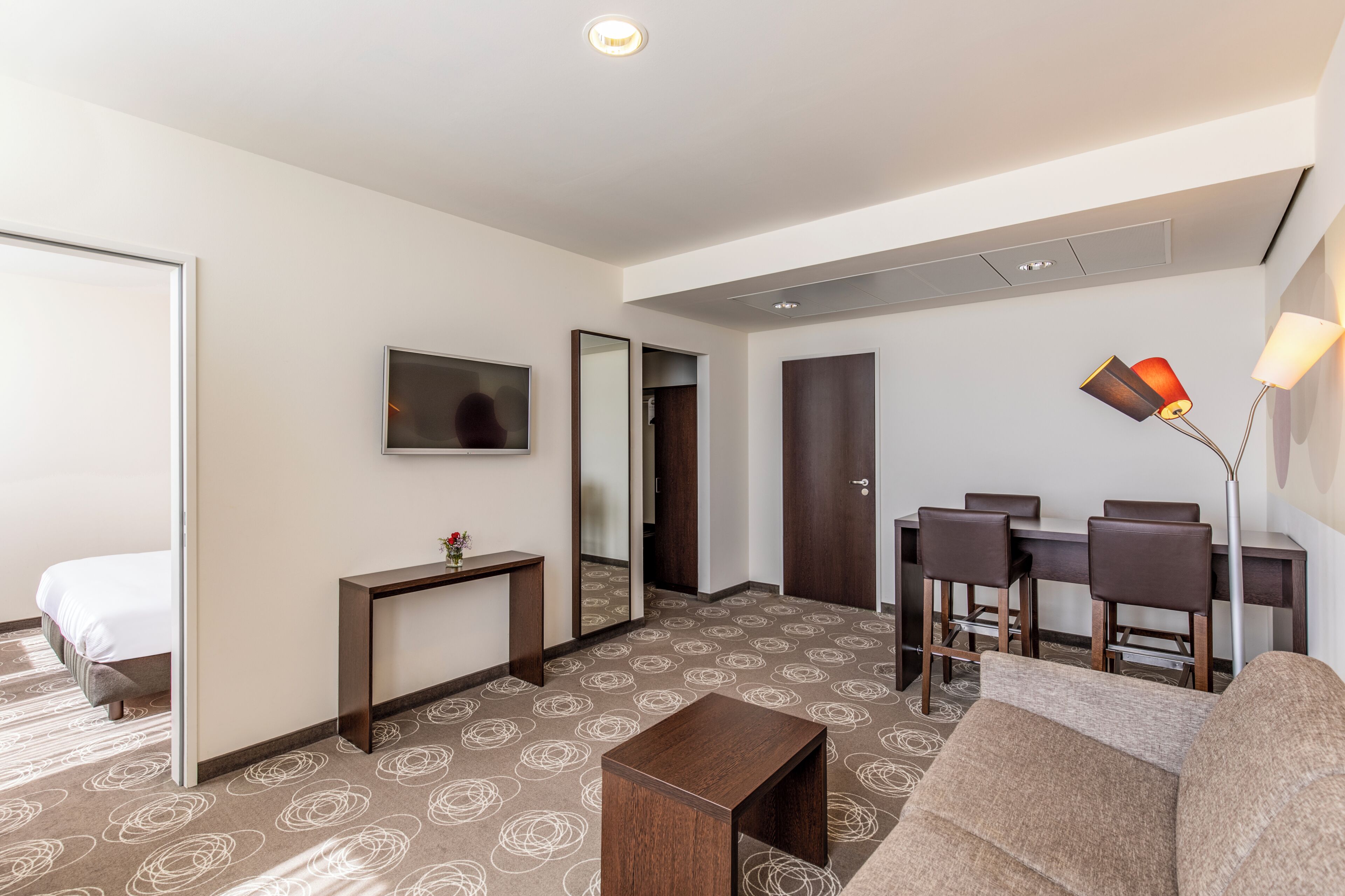 A hotel room with a bed, a desk, a chair, a sofa, a TV, and a lamp.