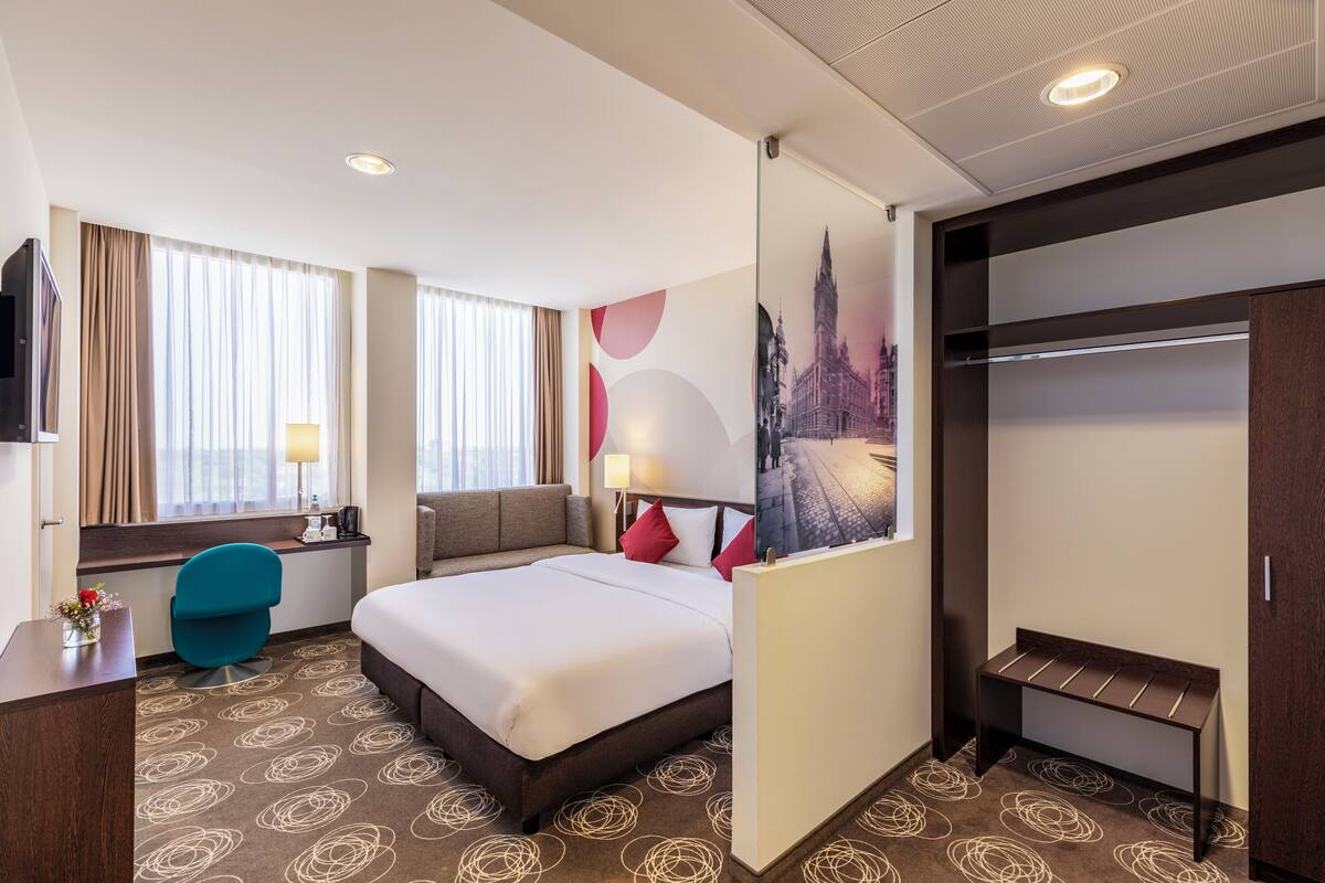 Une chambre d’hôtel comprenant un lit, un bureau, une chaise, une armoire et un mur orné d’un tableau représentant un paysage urbain.