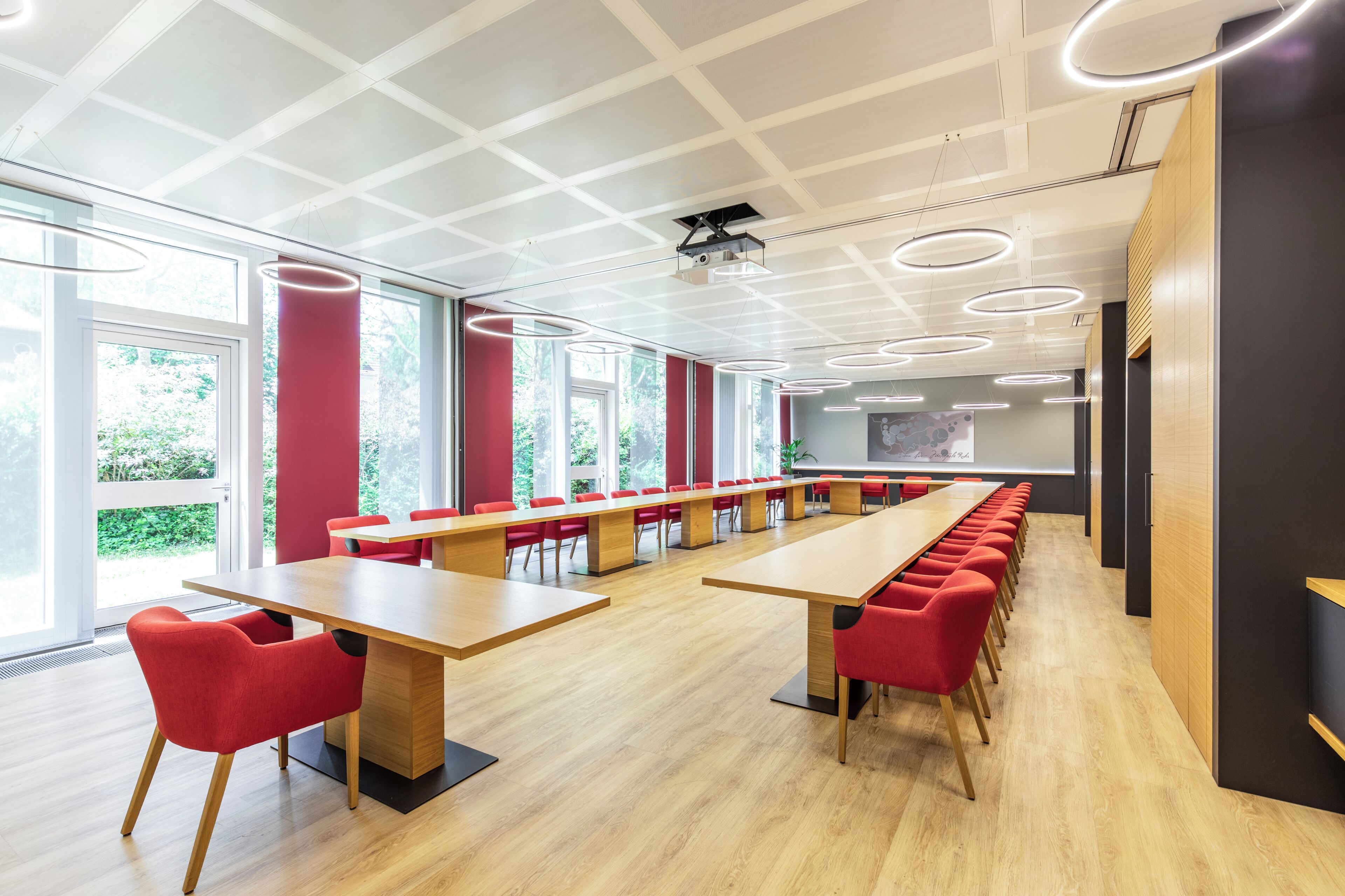 Une salle de conférence dotée de longues tables et de chaises rouges, de grandes fenêtres et d’un plafond au design moderne.