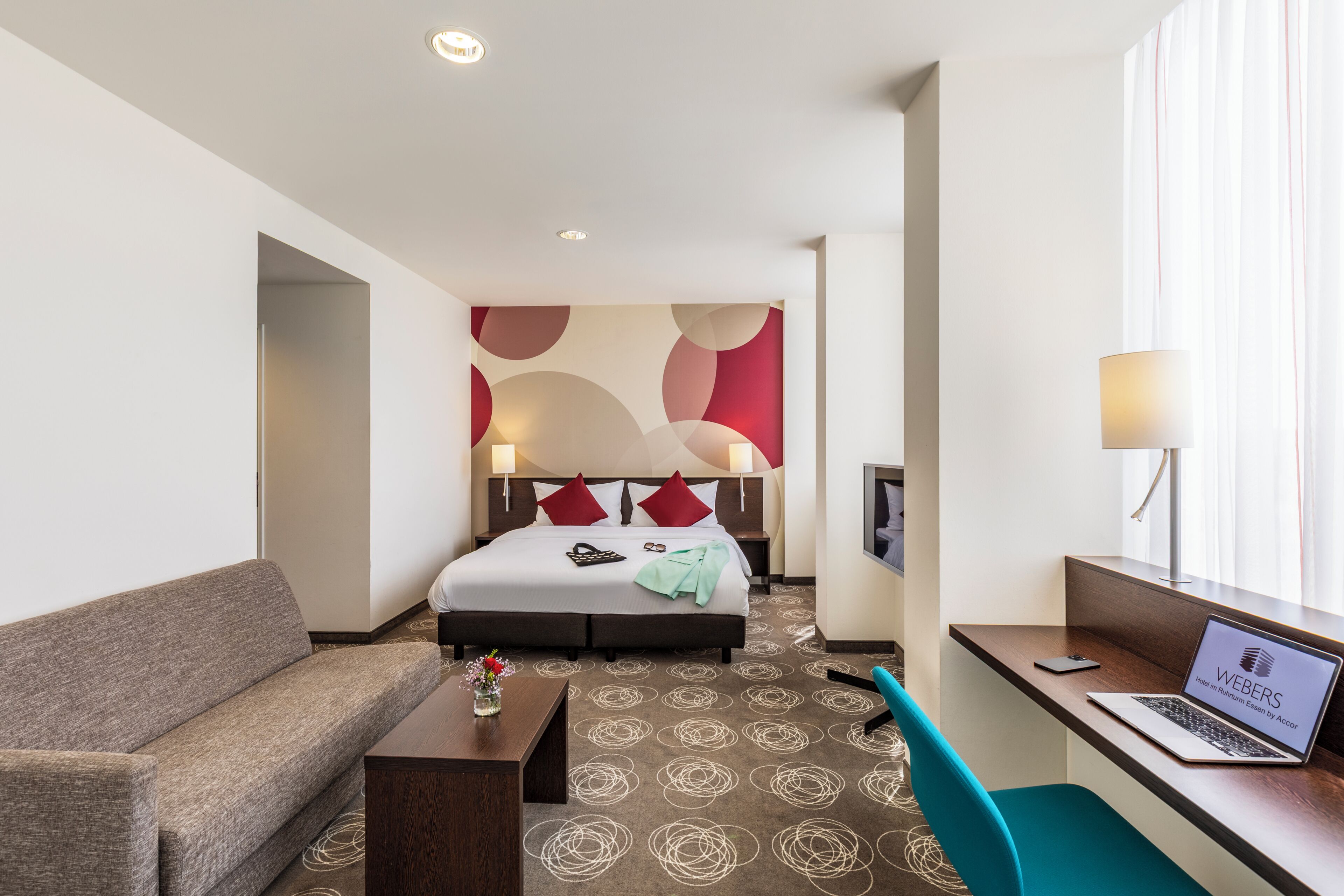 A hotel room with a bed, a desk with a laptop, a sofa, and a small table with a vase.