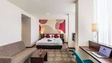 A hotel room with a bed, a desk with a laptop, a sofa, and a small table with a vase.