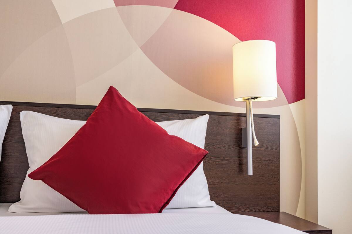 Un lit avec des oreillers blancs et rouges, une tête de lit en bois et une lampe fixée au mur.