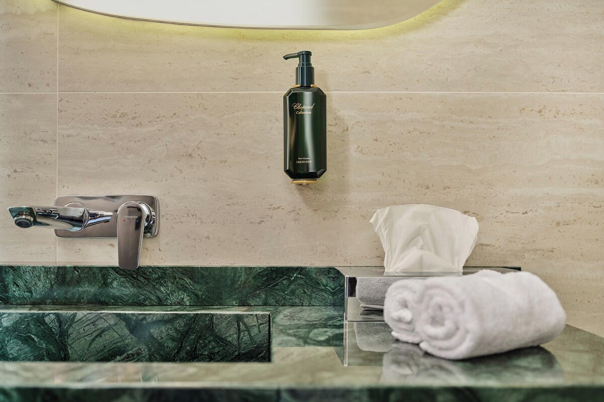 A bathroom with a marble sink, a handheld showerhead, a bottle of shampoo, and a towel.