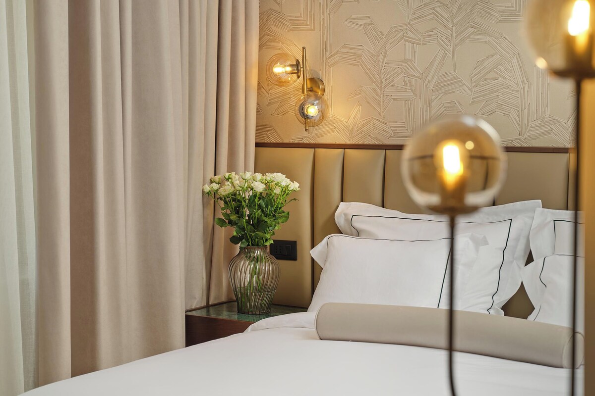 A hotel room with a bed, a nightstand with a vase of flowers, and a wall-mounted light fixture.