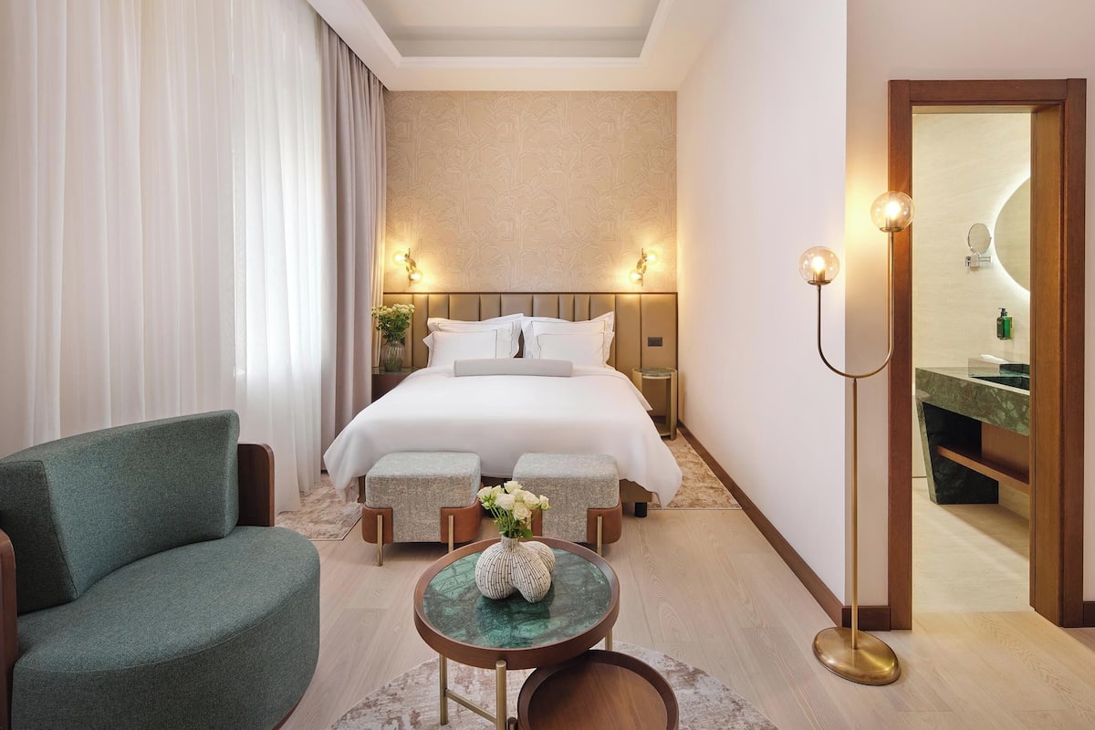 A hotel room with a bed, a green armchair, a small table with a vase, and a mirror in the corner.