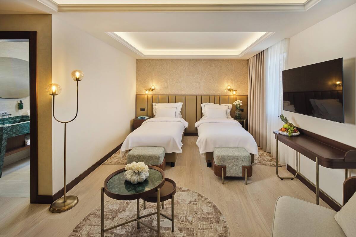 A hotel room with two beds, a TV, a small table with a vase, and a nightstand with a lamp.