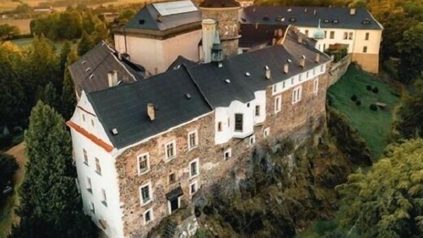 Et stort, historisk slot med flere tårne og et markant, centralt tårn, omgivet af frodig vegetation og bakker.