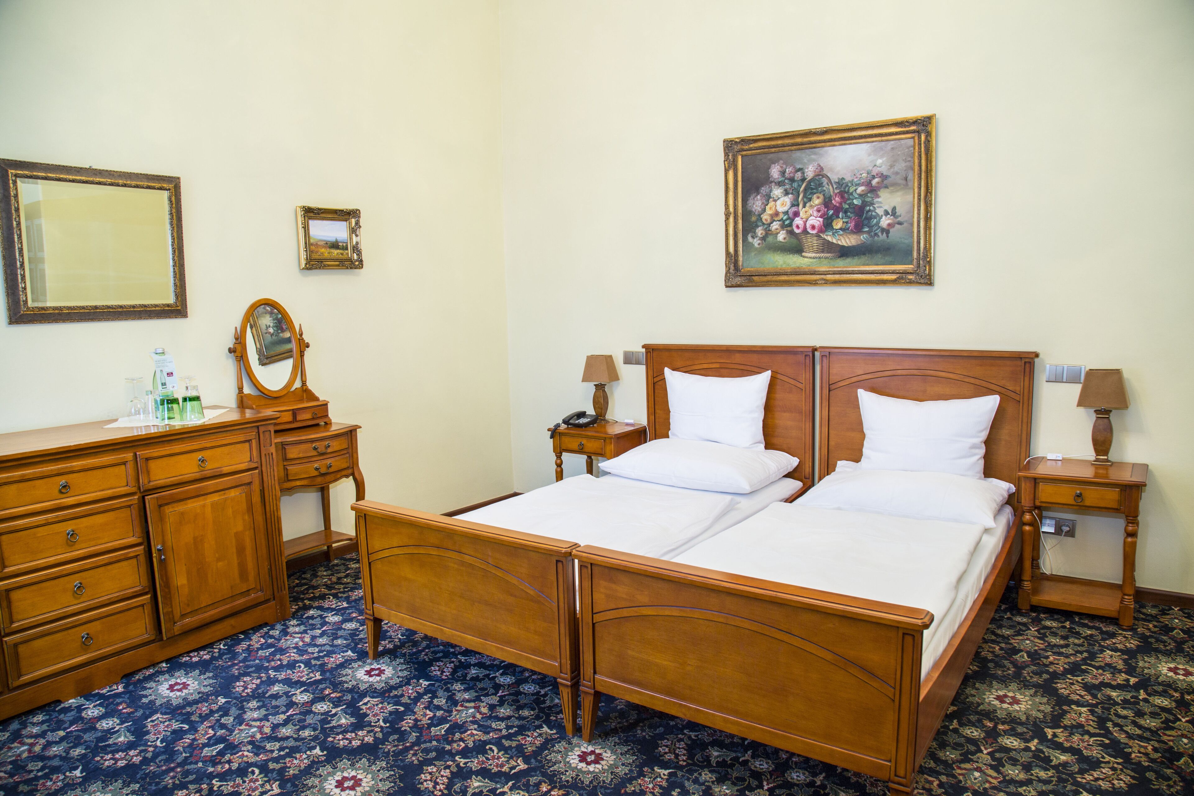 Una cama doble con cabecera de madera, una mesita de noche con lámpara, un aparador con espejo y una alfombra estampada con motivos florales.