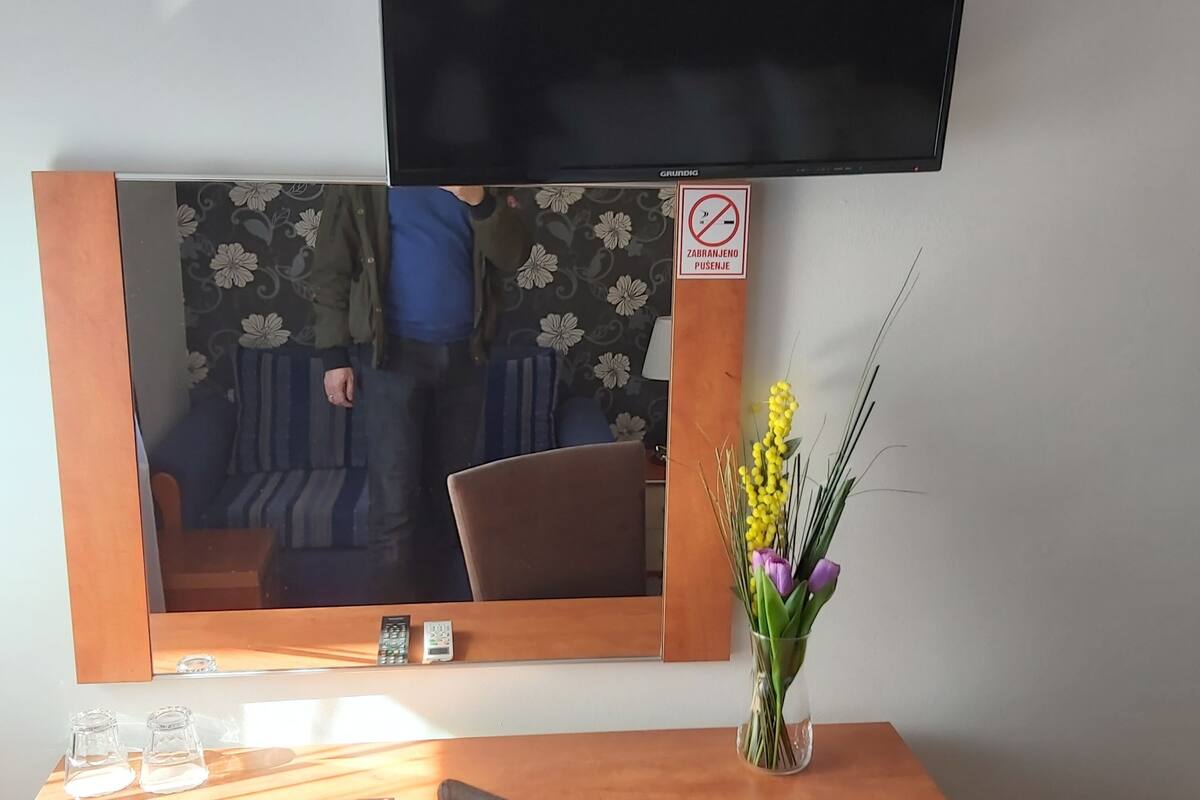 A hotel room with a wooden desk, a flat-screen TV mounted above it, a mirror reflecting part of the room, and a vase with flowers on the desk.