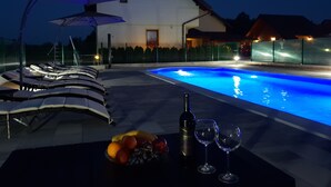 Een zwembad met ligbedden en parasols, een tafel met fruit en wijnglazen, en een gebouw op de achtergrond.