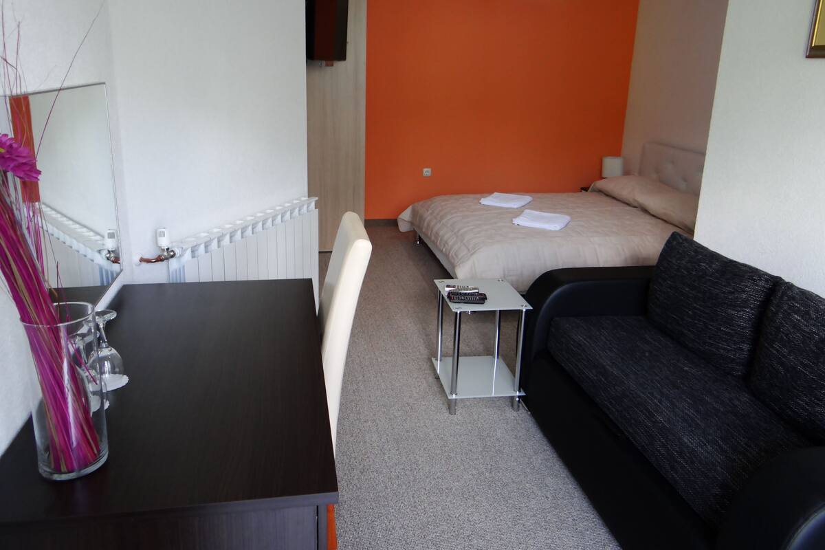 A compact hotel room with a bed, a sofa, a small table, and a mirror.