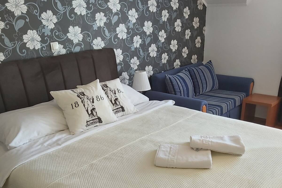 A neatly made bed with white bedding and decorative pillows, a blue sofa, and a patterned wallpaper in the background.