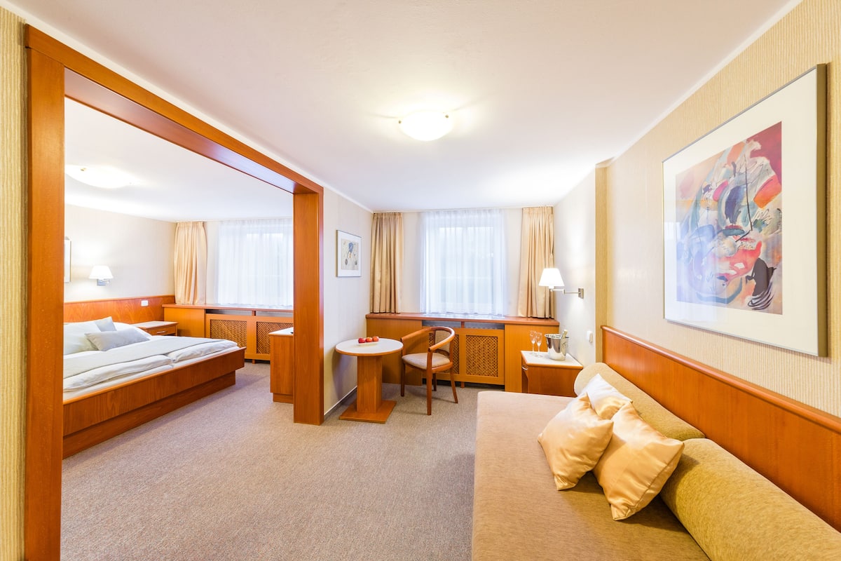 A hotel room with a bed, a sofa, a desk, and a chair. There is a painting on the wall and a window with curtains.