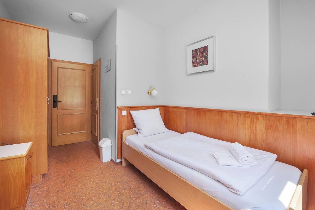 A single bed with a wooden headboard, a white pillow, and a rolled towel on the bed. There is a small trash bin next to the bed and a door leading to another room.