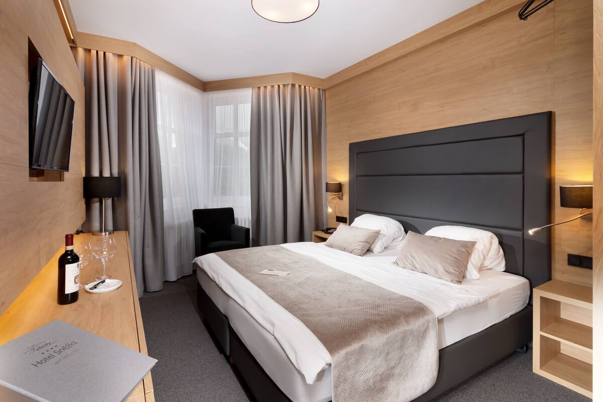 A modern hotel room with a large bed, a flat-screen TV, a desk with a wine bottle and glasses, and a nightstand with a lamp.
