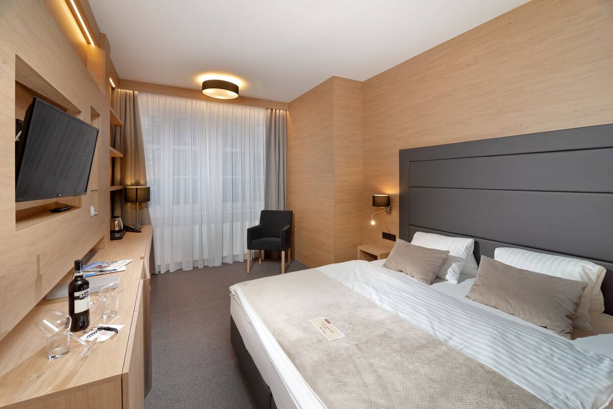 A modern hotel room with a large bed, a flat-screen TV mounted on the wall, a desk with a bottle of wine and glasses, and a chair.