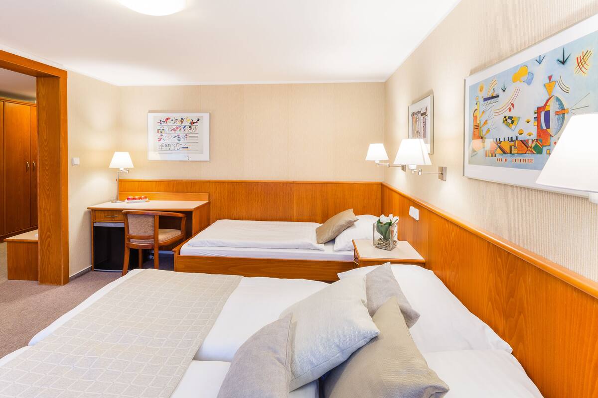 A hotel room with two beds, a desk, and a chair. There are wall-mounted lamps and a painting on the wall.