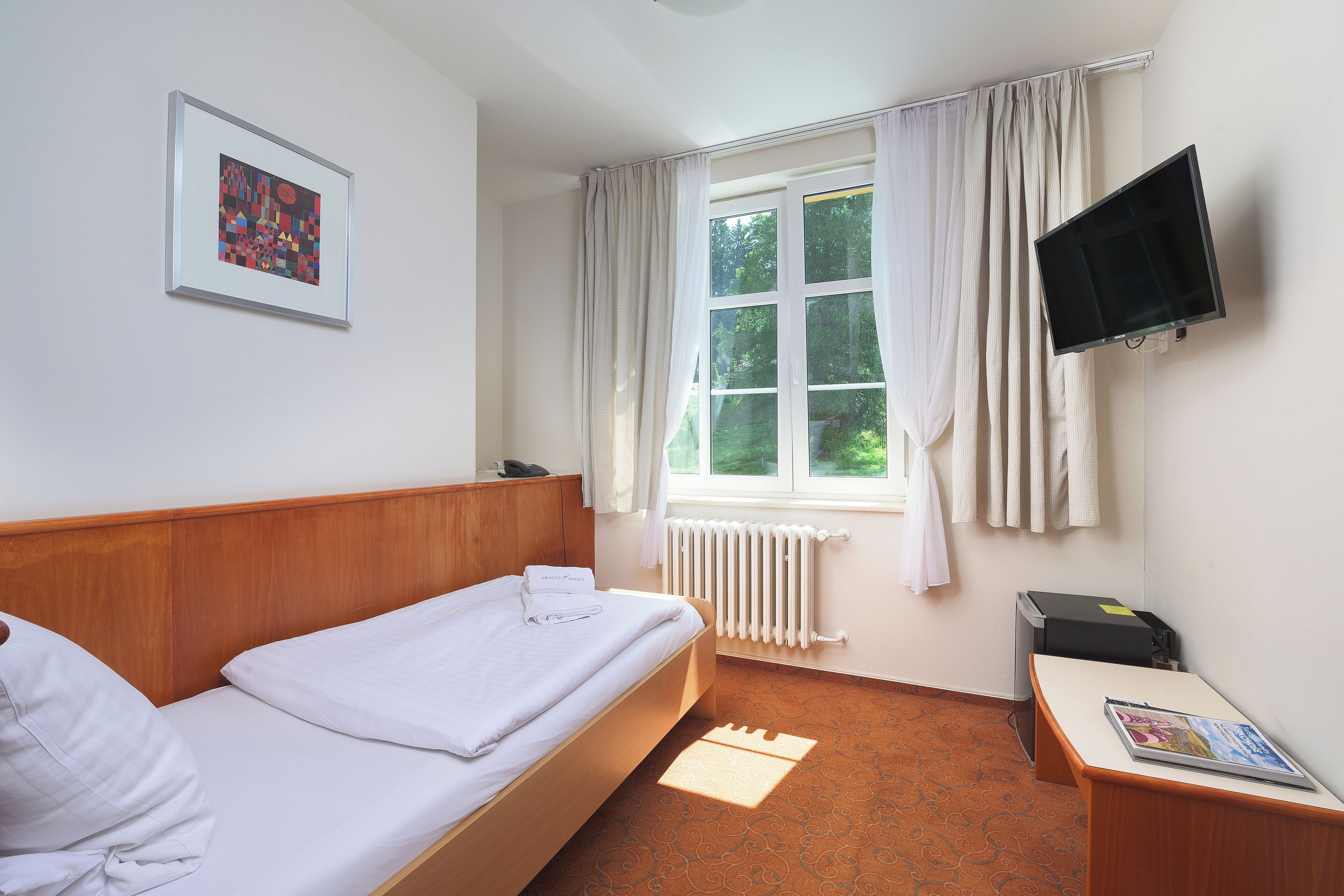 Habitación de hotel con una cama individual, un televisor montado, un radiador y una ventana con cortinas.