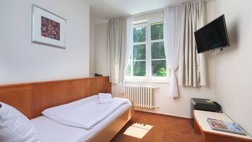 Habitación de hotel con una cama individual, un televisor montado, un radiador y una ventana con cortinas.
