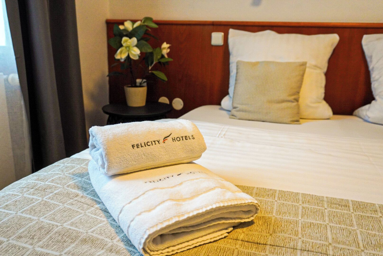 A neatly made bed with a folded towel on the side, a bedside table with a potted plant, and a wall-mounted light switch.