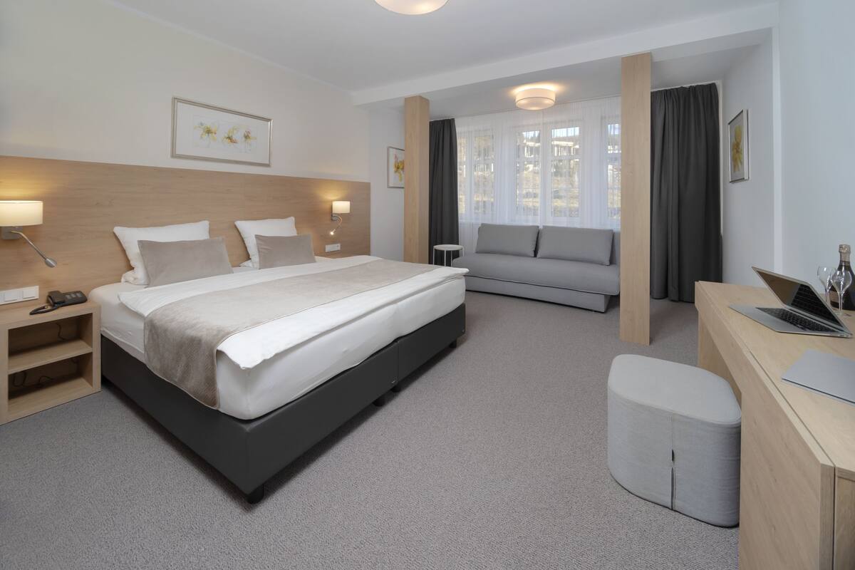 A modern hotel room with a large bed, a sofa, a desk with a laptop, and a small ottoman.