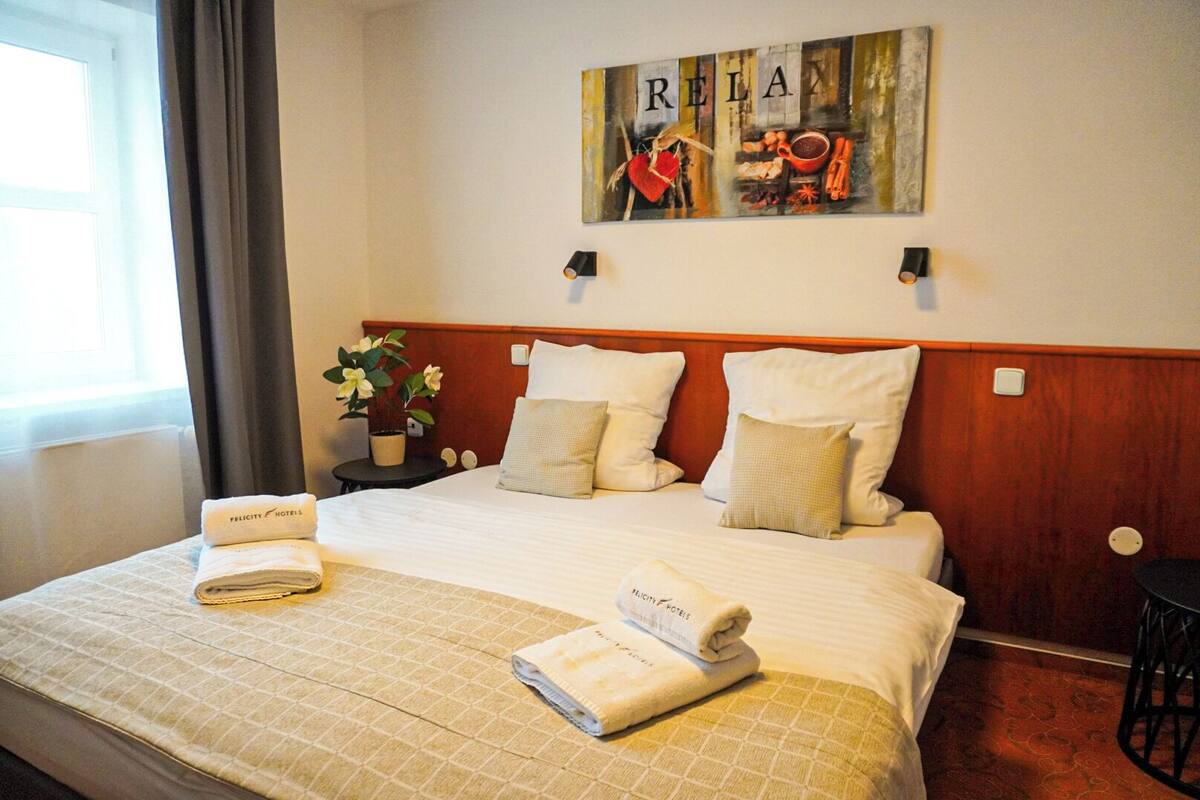 A hotel room with a bed, pillows, a nightstand with a plant, and a painting on the wall.