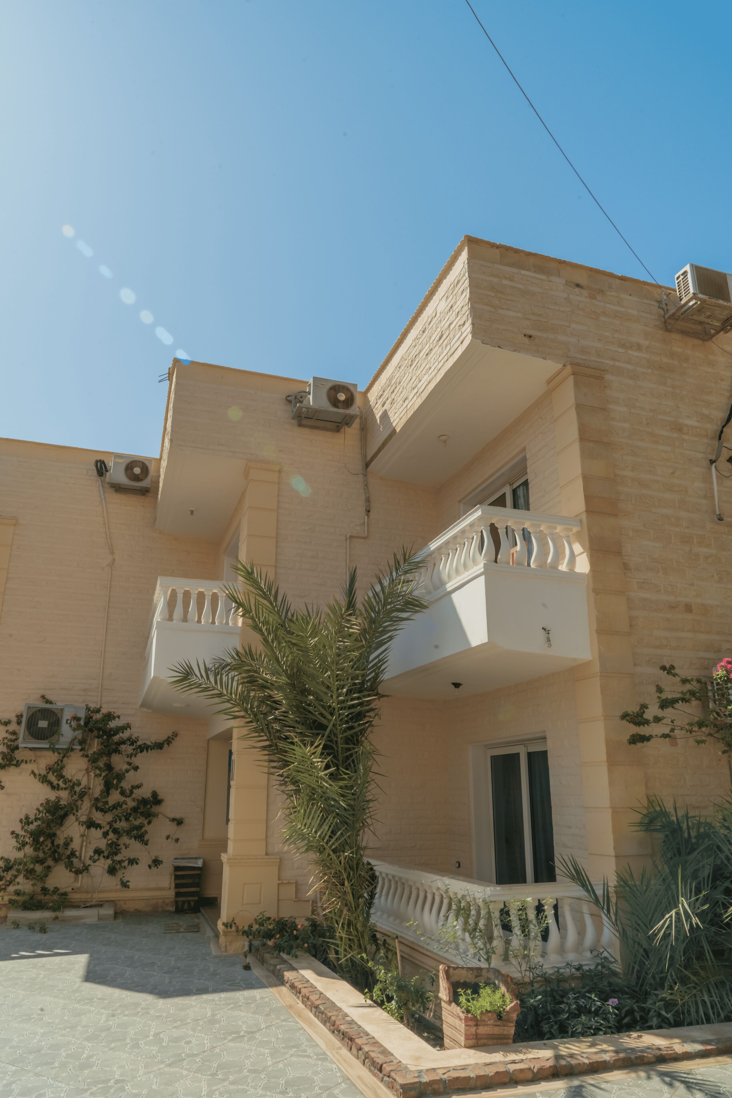 A two-story building with a balcony on the second floor, a small garden area with plants, and a clear sky.