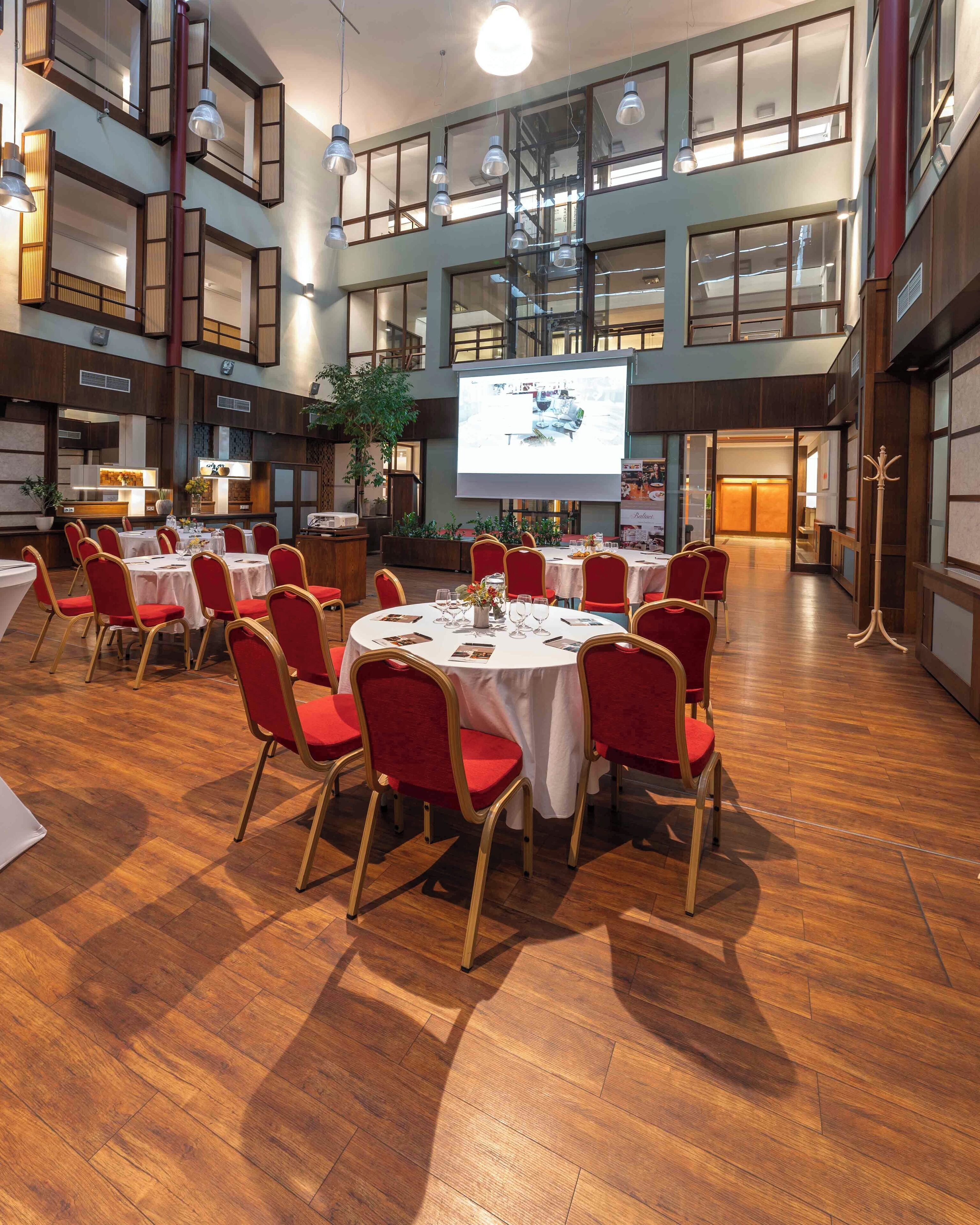 A spacious hall with multiple round tables and red chairs, wooden floors, and a large screen displaying a presentation.