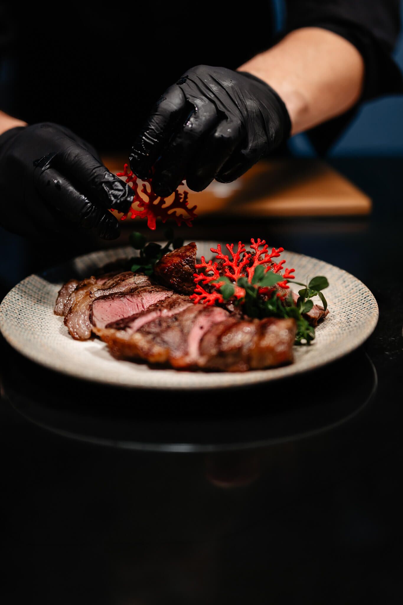 A plate of sliced meat with garnish, a person wearing black gloves, and a dark background.