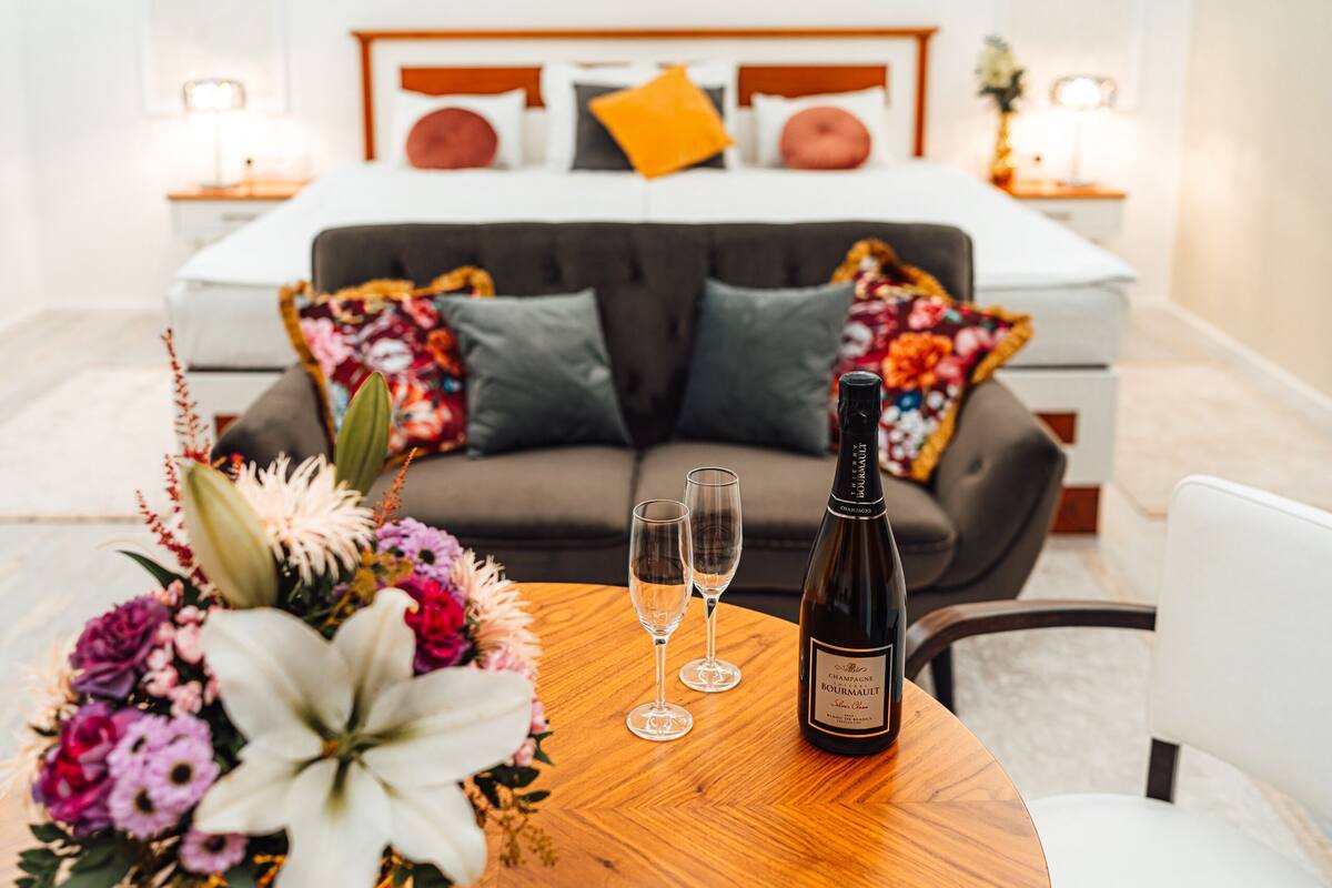 A room with a bed, a wooden table with a bottle and two glasses, and a floral arrangement.