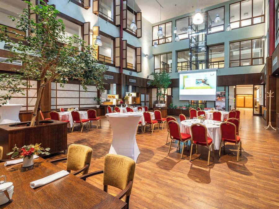 Een ruime vergaderzaal met meerdere tafels en stoelen, een groot scherm voor presentaties en een boom in het midden.