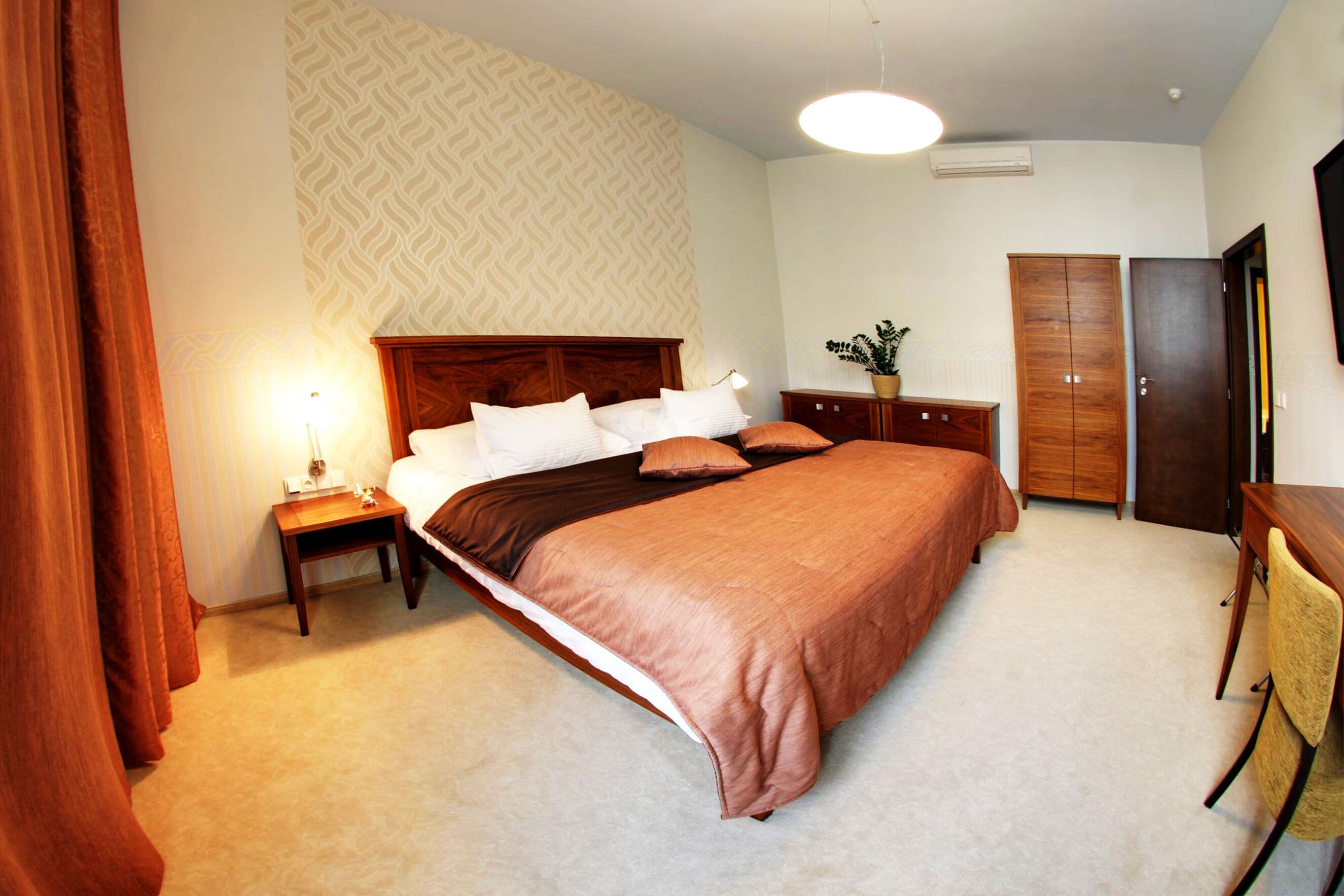 A neatly arranged bedroom with a large bed, a bedside table with a lamp, a wardrobe, and a desk with a chair.