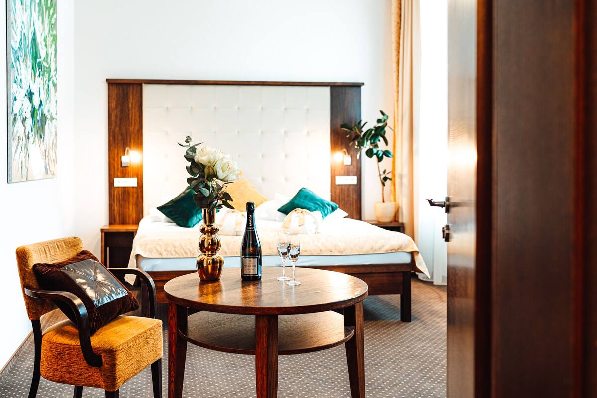 A hotel room with a bed, a round wooden table with a bottle and glasses, a chair with a cushion, and a painting on the wall.