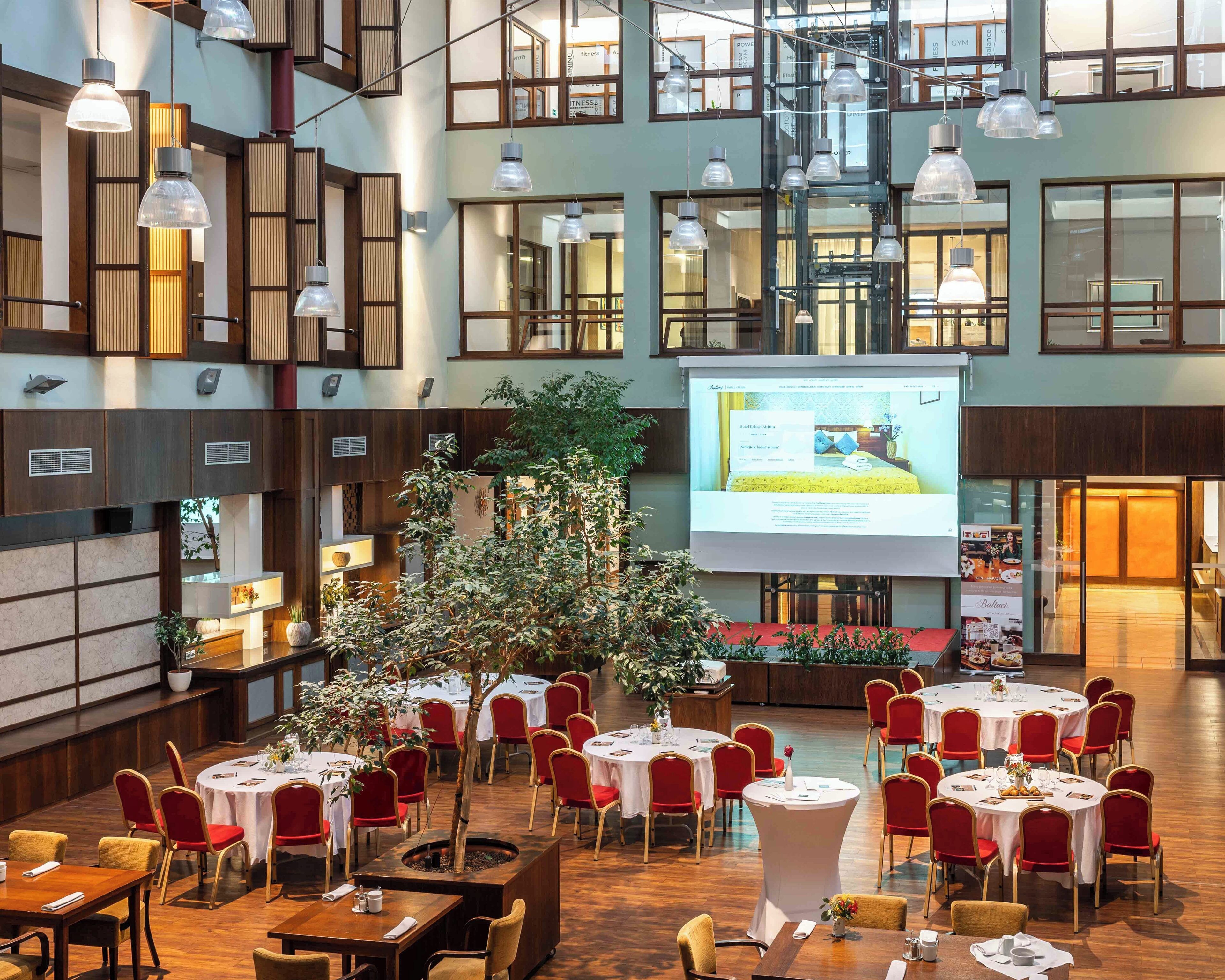 A spacious indoor venue with multiple dining tables and red chairs, a central tree, and a projection screen displaying a room.