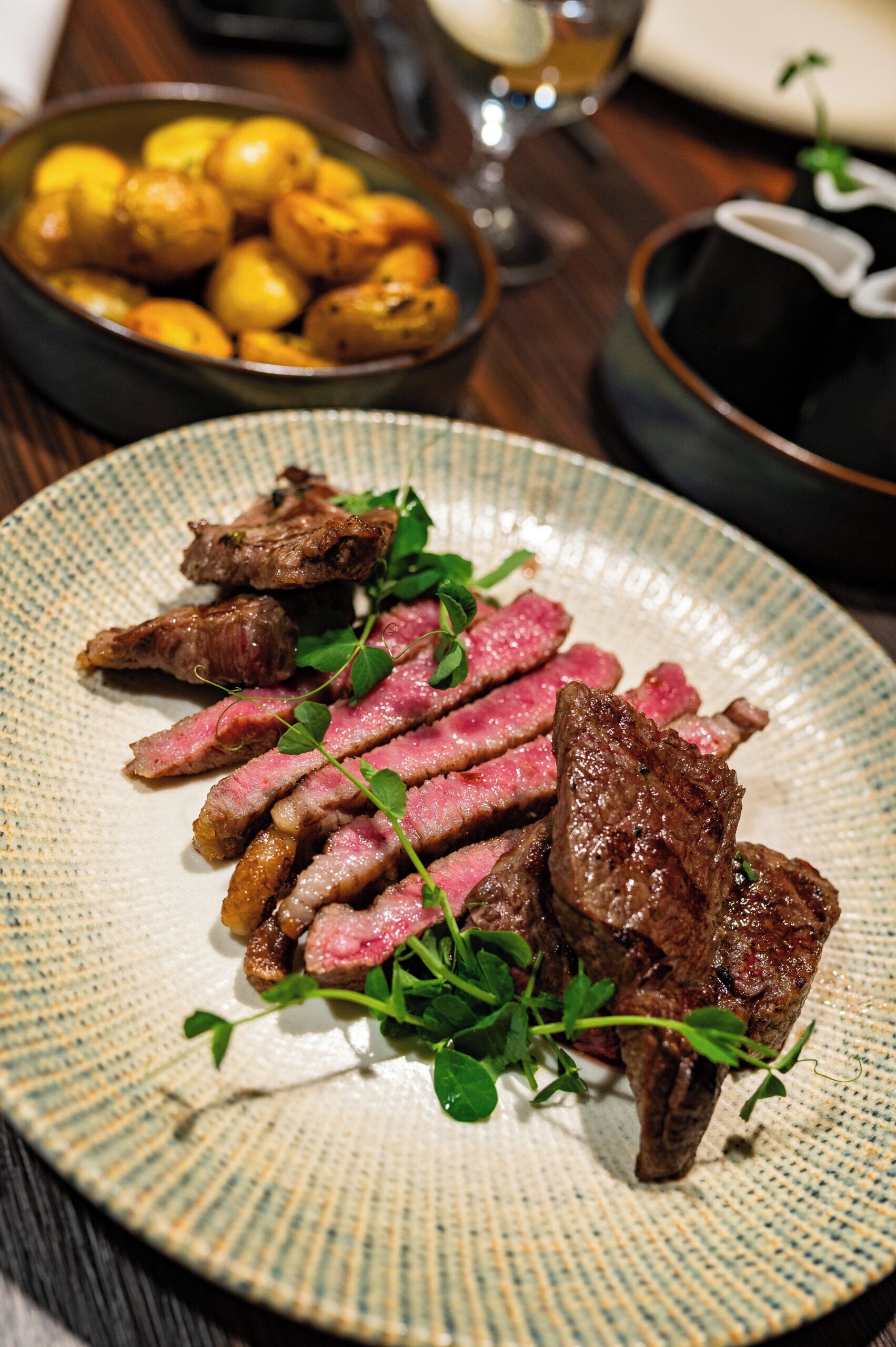 A plate of sliced meat with greens, a bowl of potatoes, and a glass of wine on a wooden table.
