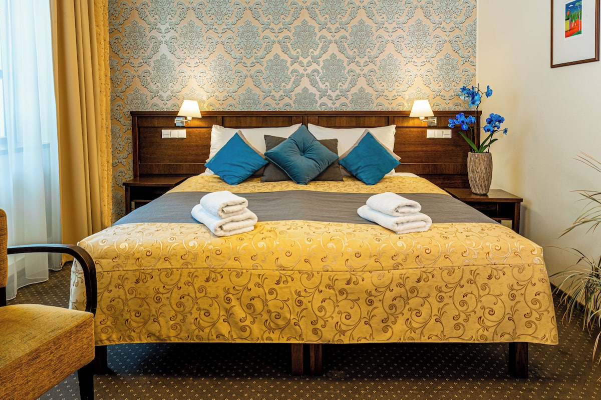 A hotel room with a bed, two bedside tables, a chair, and a wall with a patterned wallpaper.