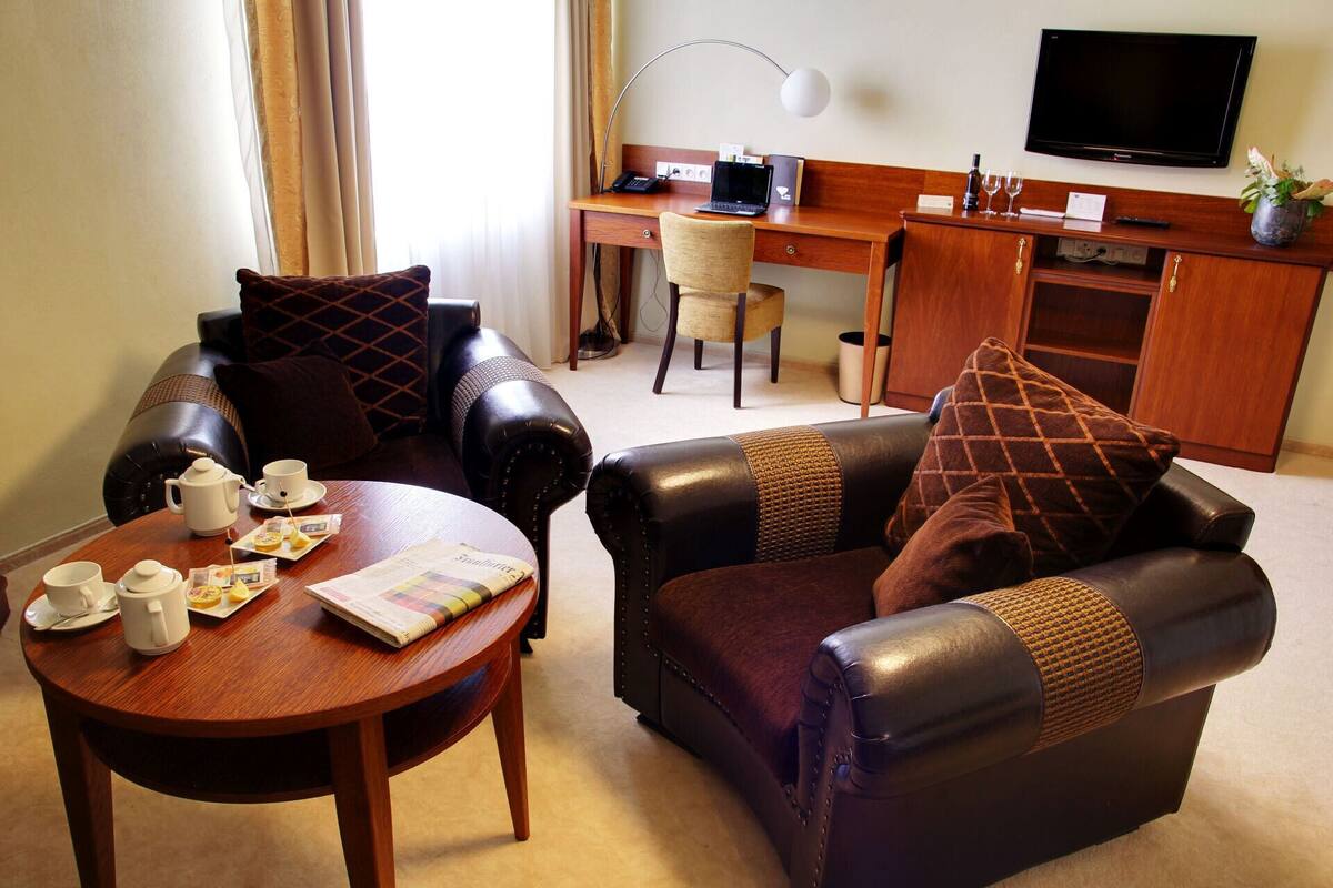 A hotel room with a leather sofa, a wooden coffee table with a newspaper, a desk with a laptop, a television, and a small plant.