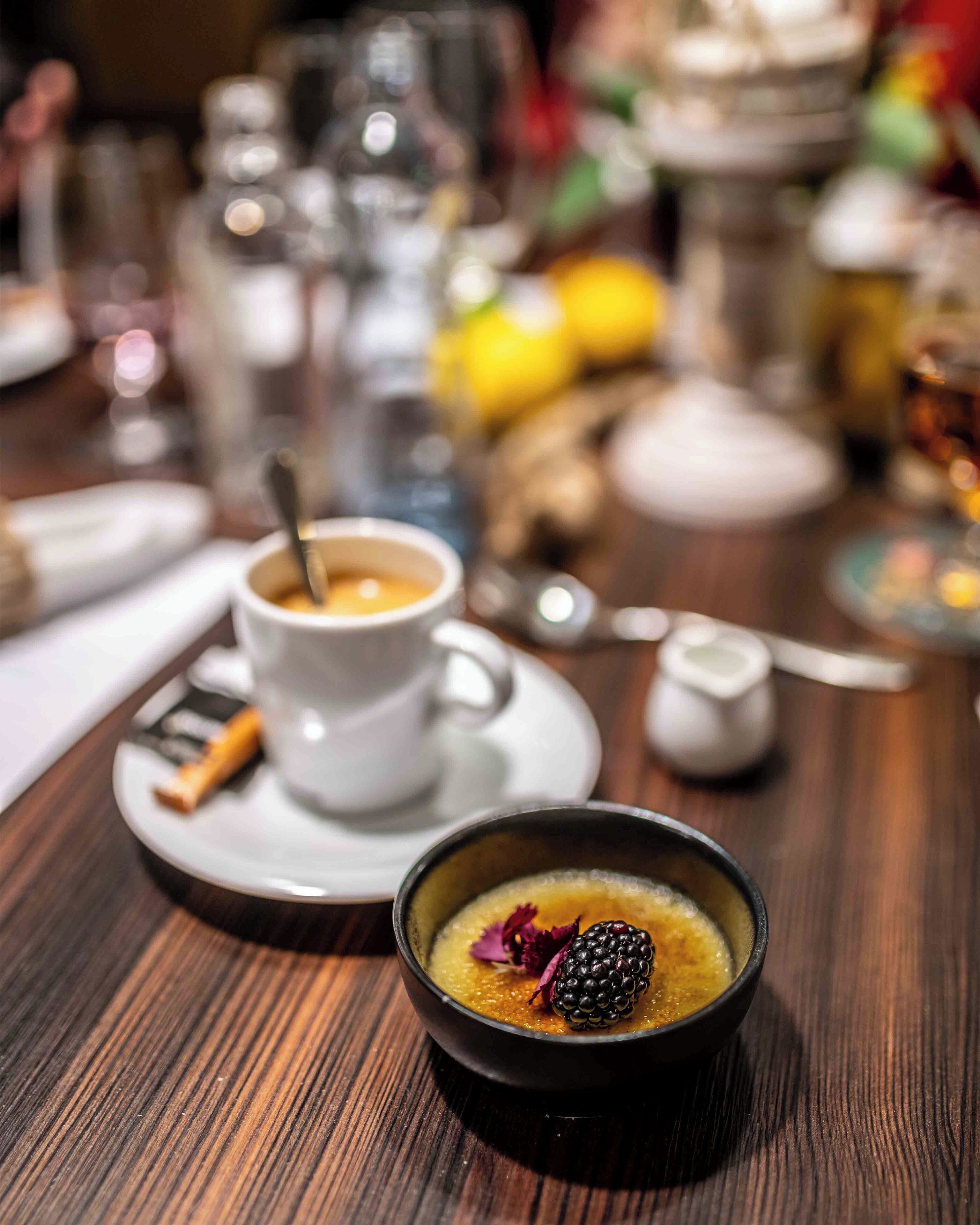 A dining table with a cup of coffee, a bowl of soup garnished with a blackberry, and a spoon.