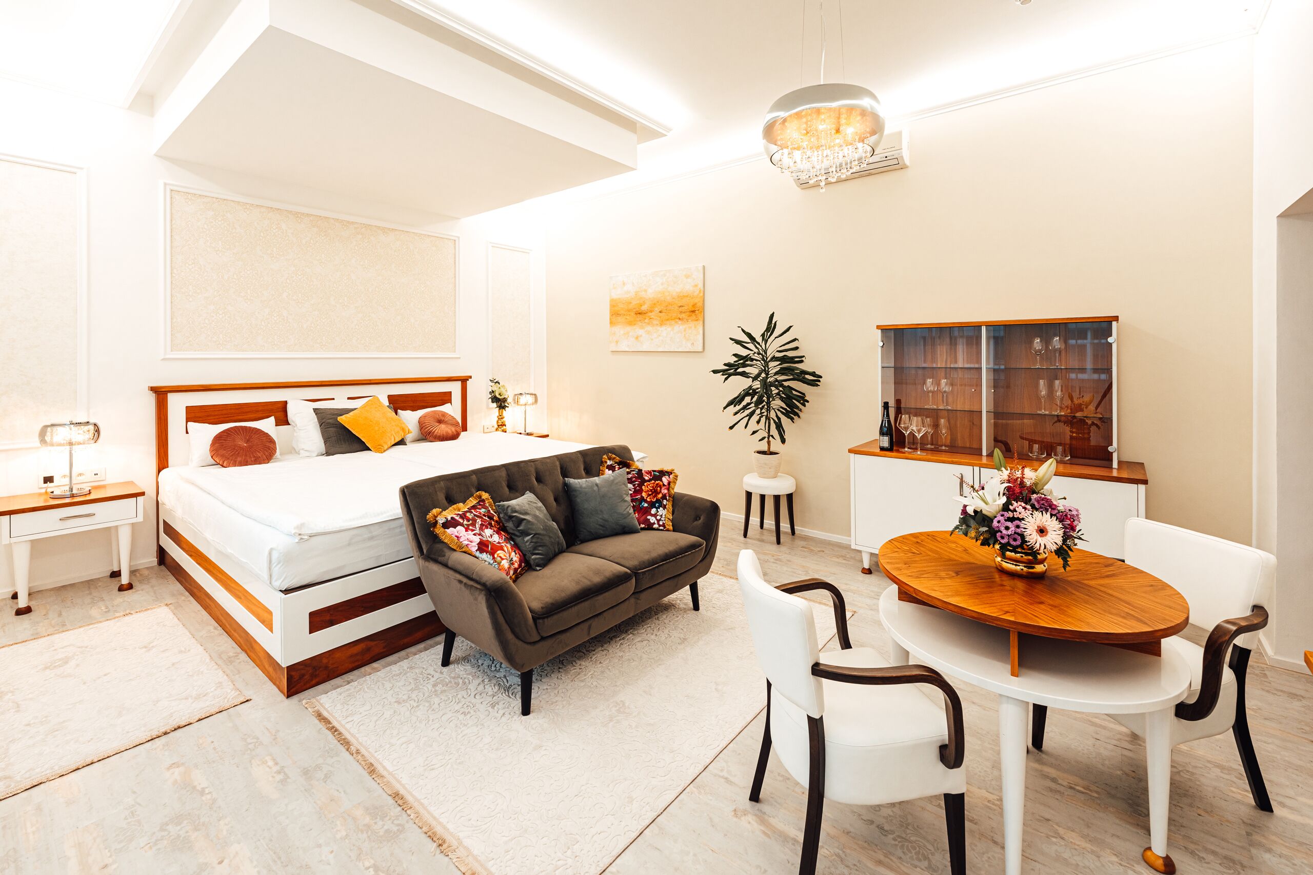 A modern bedroom with a bed, a sofa, a dining table with chairs, a small side table, and a decorative plant.