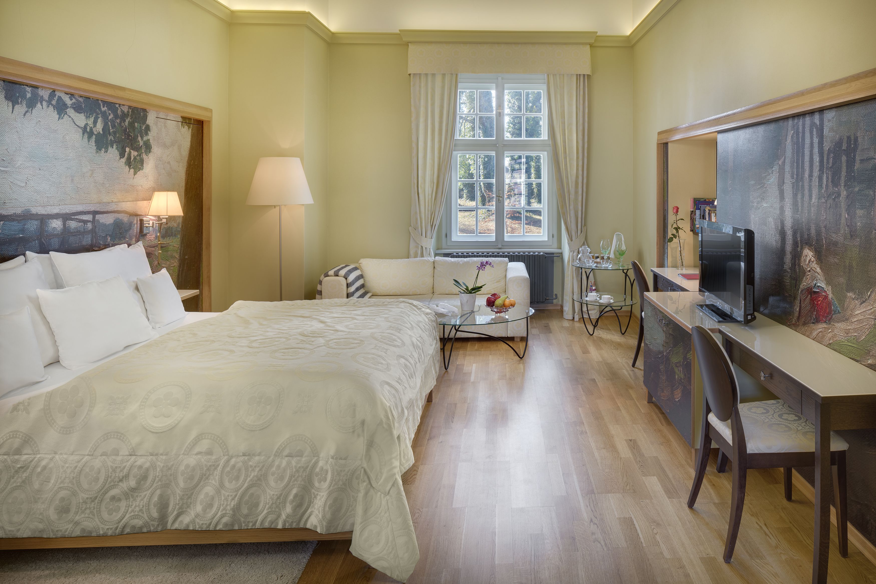 Een hotelkamer met een groot bed, een bureau met een computer, een stoel, een klein tafeltje met een bloemenvaas en een raam met gordijnen.