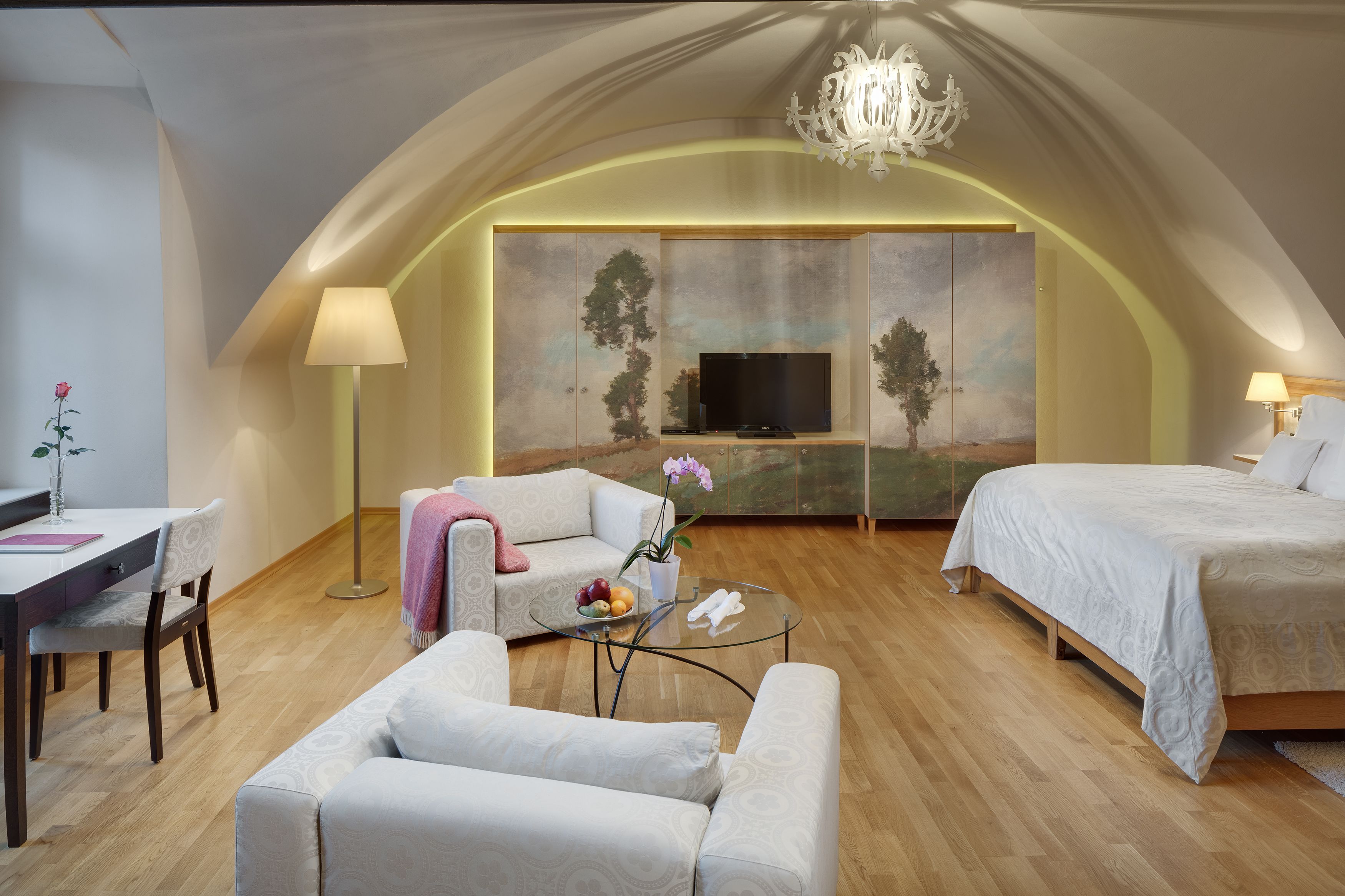 Ein modernes, gut beleuchtetes Zimmer mit gewölbter Decke, einem Fernseher, einer Couch, einem Bett und einer Essecke mit Tisch und Stühlen.