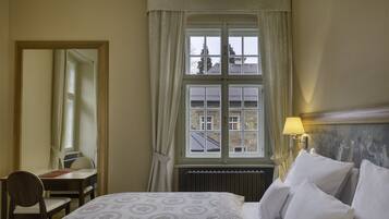 A hotel room with a bed, a desk with a chair, a window with curtains, and a lamp.